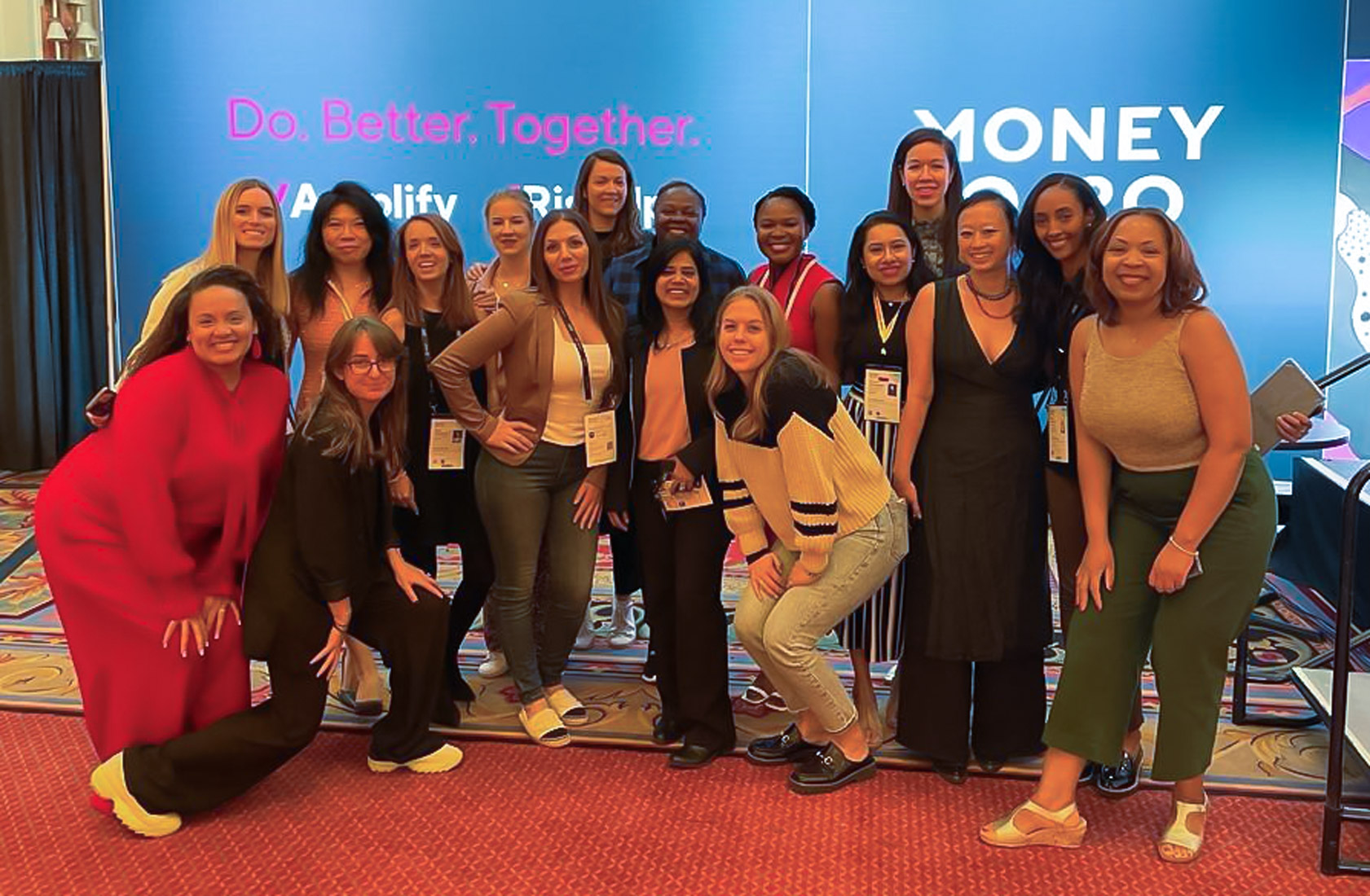 A group photograph of the Money 20/20 RiseUp cohort, made up of 17 women. They are standing in front of a Money 20/20 banner in a large ball room.