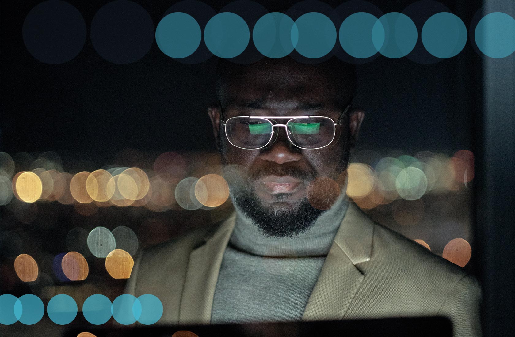 African American man is looking at a screen which is reflecting on his gold-rimmed glasses. He's standing in front of a large window that looks out over a city at night.